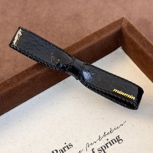 Detail of a black faux leather bow hair clip with a 3D golden minmin metal letter logo