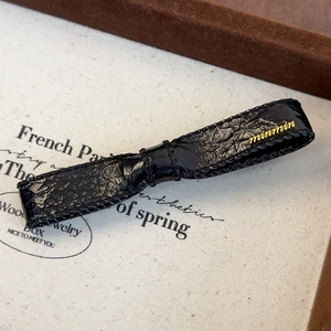 Detail of black leather bow hair clip with a gold logo on a textured canvas base