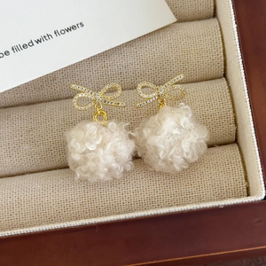 Close-up of fluffy cream plush pompom earrings with sparkling bowknot studs in a gift box