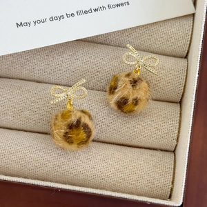 Close-up of golden bow earrings with micro-set zircons and soft leopard print pompom