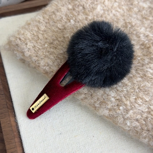 Détail d'une pince à cheveux en velours bordeaux avec pompon noir et étiquette dorée