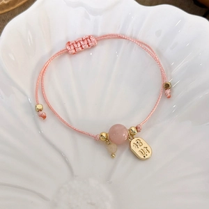 Close-up of pink woven bracelet with lucky gold pendant and beads on white shell tray