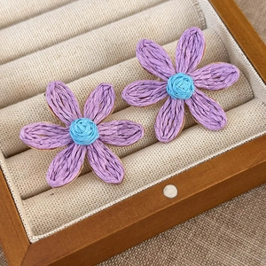 Detailed close-up of woven raffia sunflower earrings in purple and blue on linen
