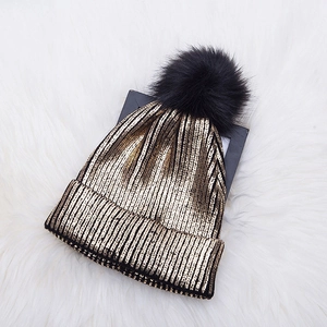 Golden knit beanie hat with black faux fur pom-pom on white faux fur background