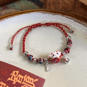 Macro detail of Maneki Neko charm bracelet with red woven cord and ceramic beads on a tray
