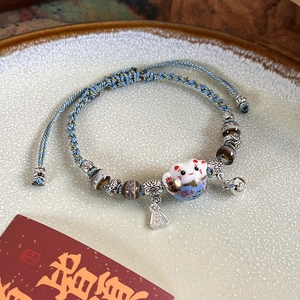 Detail of hand-woven cat charm bracelet with ceramic lucky cat and gold bell on a plate