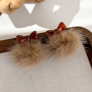 Dettaglio orecchini a pompon marroni e fiocco in resina ambrata su vassoio di lino grigio