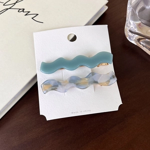 Detail of two wavy acetate hair clips in teal and marble blue on a white display card