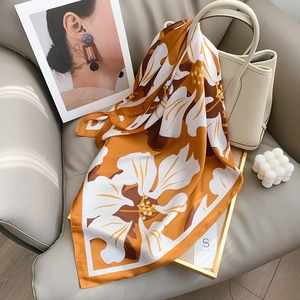 Elegant orange silk scarf with white floral print styled on a leather sofa with luxury bag
