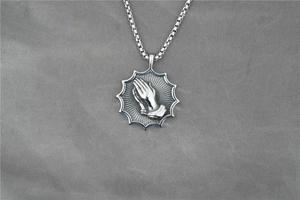 Détail macro du pendentif mains en prière argenté avec chaîne box sur tissu gris texturé