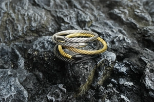 Detailed view of woven cable open bangles in gold, silver, and black on a grey rock