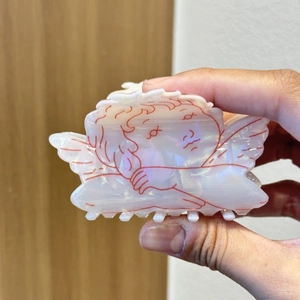 Detail of pearlescent angel wing hair claw clip with red cupid motifs held in hand