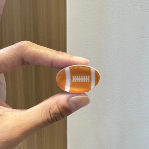 Dettaglio di un mini fermaglio per capelli a forma di football mostrato sul dito