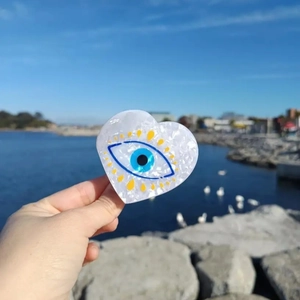 Handheld heart-shaped Evil Eye hair claw clip with pearlescent finish against ocean background