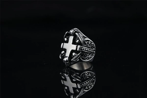 Detalle de anillo gótico de acero con cruz y calaveras sobre fondo negro con reflejo