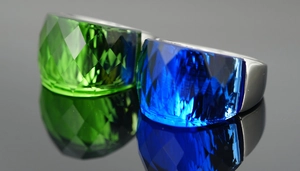 Detail of blue and green faceted gemstones set in heavy metal rings on a reflective surface