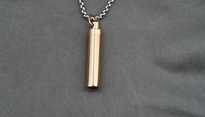 Close-up of rose gold cylindrical urn pendant and silver steel chain on grey fabric