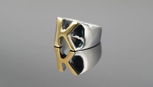 Vue macro détaillée de la bague argentée large avec une lettre K dorée sur surface miroir