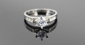 Macro shot of silver six-prong diamond ring with side stones on a reflective black surface