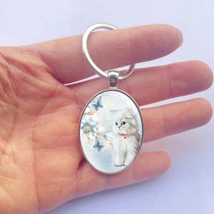 Porte-clés ovale en métal avec chat blanc, fleurs et papillons tenu dans la main