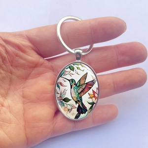 Handheld view of a polished metal oval keychain featuring a colorful hummingbird pattern