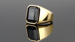Bague en acier doré poli avec pierre noire rectangulaire sur fond réfléchissant sombre