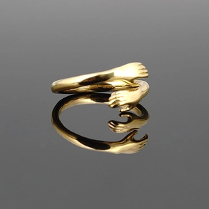 Polished gold embrace ring with hand-shaped hug design on a reflective grey surface