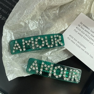 Detail of dark green resin hair clip with AMOUR rhinestone lettering on paper background