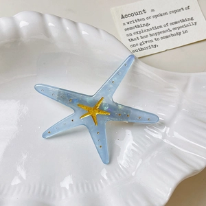 Close-up of translucent blue starfish hair clip with gold accents in a white shell tray