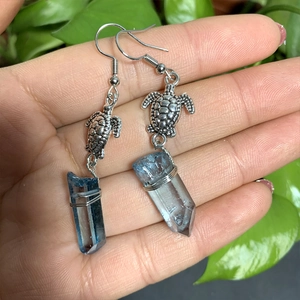 Detail of silver turtle earrings with sea blue crystal stones held in hand against foliage