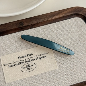 Close-up of a dark green acetate hair clip with gold lettering on a beige linen base