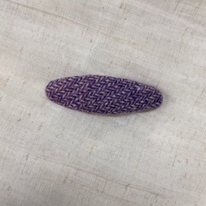 Close-up of purple herringbone tweed oval hair clip on textured beige linen fabric
