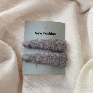 Close-up of a pair of gray plush triangular BB hair clips on a soft beige fabric background