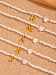 Detailansicht von fünf weißen Perlenketten mit goldenen Buchstabenanhängern auf Sand