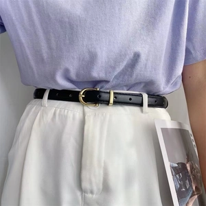 Détail d'une fine ceinture en cuir noir avec boucle fer à cheval dorée sur pantalon blanc