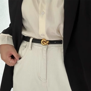 Detail showing a thin black leather belt with a golden knot buckle on beige trousers