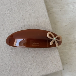 Close-up of a brown acetate hair clip with a small beige bow and gold lettering