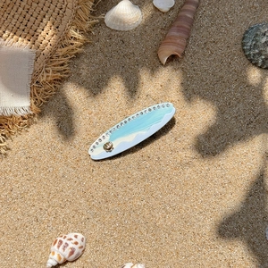 Surfboard hair clip with blue ocean waves and gold shell decor on sandy beach