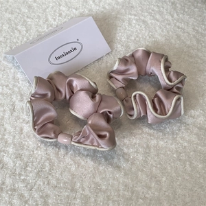 Detailansicht von zwei rosa Satin-Haargummis mit weißer Paspelierung auf weichem Plüschgrund