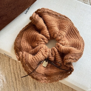 Macro detail of caramel knit bow scrunchie with gold tag on a white book surface