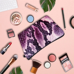 Flat lay of snakeskin makeup bag with brushes and cosmetics on light pink background