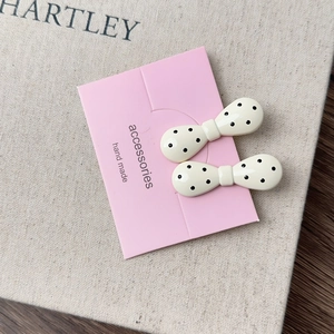 Close-up of two cream bow hair clips with black polka dots on a pink display card