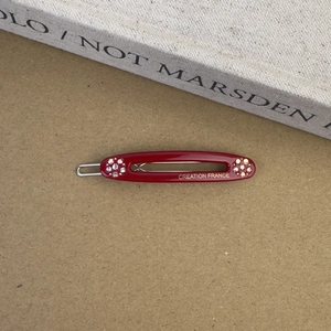 Détail de la barrette ovale en acétate bordeaux avec strass et lettrage doré élégant