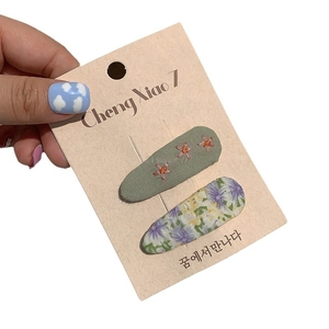 Handheld set of two floral embroidery hair clips on a card featuring green and purple patterns