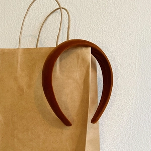Velvet padded headband in burnt orange hanging on a paper bag with a minimalist backdrop
