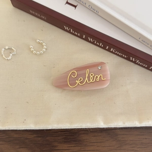 Close-up of pink marble acetate hair clip with gold script lettering and pearl accent