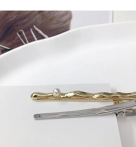 Close-up of gold wave-textured pearl hairpin and silver metal duckbill clip on white stand