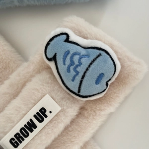 Dettaglio sciarpa in peluche beige con toppa a pesce blu ed etichetta GROW UP cucita