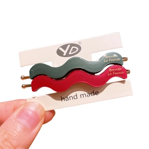 Handheld view of wave-shaped matte hair clips in dark green and deep red with gold accents