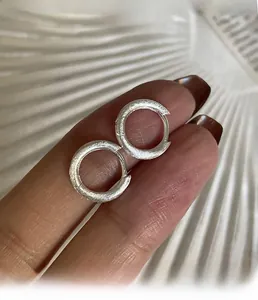 Dettaglio degli orecchini a cerchio in metallo spazzolato argento tenuti a mano su sfondo bianco